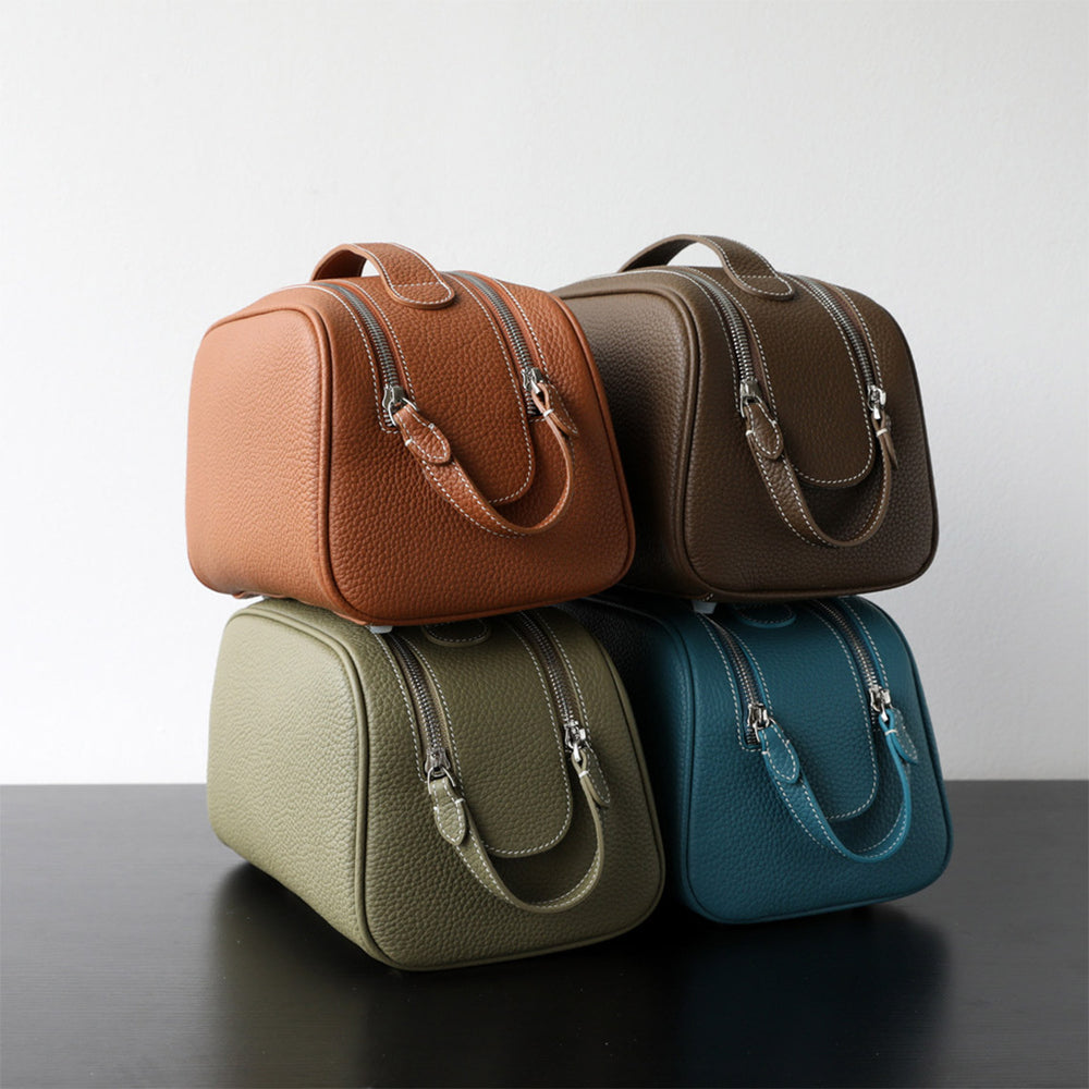 Four leather makeup bags in taupe, green, blue, and gold brown stacked on a reflective surface with a neutral background.